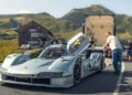 A silver Porsche 963 RSP race car with its door open is parked on a mountain road as several people stand nearby and a FAT Mankei trailer is visible behind the car.