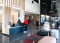 A person in a red jacket stands at a modern reception desk in a spacious, well-lit lobby at the Porsche Experience Center, with seating and a room labeled "SIMLAB SIMULATOR" visible in the background.