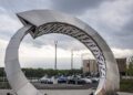 A large metallic spiral sculpture stands on a paved area outside the Porsche Experience Toronto Experience Center, with modern buildings, parked cars, and cloudy skies in the background.
