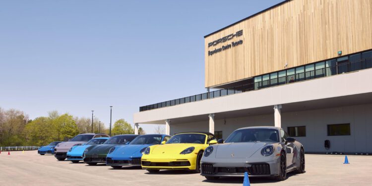 A lineup of six Porsche sports cars in various colors parked outside the Porsche Experience Center Toronto on a sunny day.