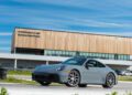 A gray Porsche sports car is parked on a track in front of the Toronto Experience Center building under a blue sky.