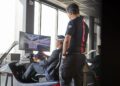 Two people are using a racing simulator at the Toronto Experience Center; one is seated holding a Porsche steering wheel, while the other stands nearby, both focused on a racing game displayed on a large screen.