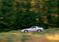 A silver Porsche sports car, painted in one of its rare colors, driving quickly past a blurred background of green trees and grass.