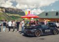 A group of people stand near several Porsche sports cars, including a 963 RSP, parked outdoors by FAT Mankei, with mountains and a wooden building in the background under a partly cloudy sky.
