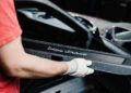 A person wearing white gloves holds a Porsche Sonderwunsch car part labeled "Porsche Lefkesenberg" next to an open car door, possibly for a 911 GT3 Touring in Luxembourg.