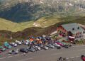 A group of sports cars, including a Porsche 963 RSP, and people gather near the FAT Mankei mountain lodge along a winding road, surrounded by green hills and high-altitude scenery.