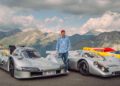 A person stands between two silver Porsche race cars, including the 963 RSP, parked outdoors with mountains and a partly cloudy sky in the background at FAT Mankei.