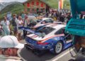 A group of people stands near several parked Porsche race cars, including a 963 RSP from FAT Mankei, at a mountain road event with a wooden building and hills in the background.