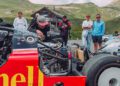 A group of people observe a mechanic working on the exposed engine of a Porsche 963 RSP with a Shell logo, surrounded by other cars near the FAT Mankei in a mountainous area.