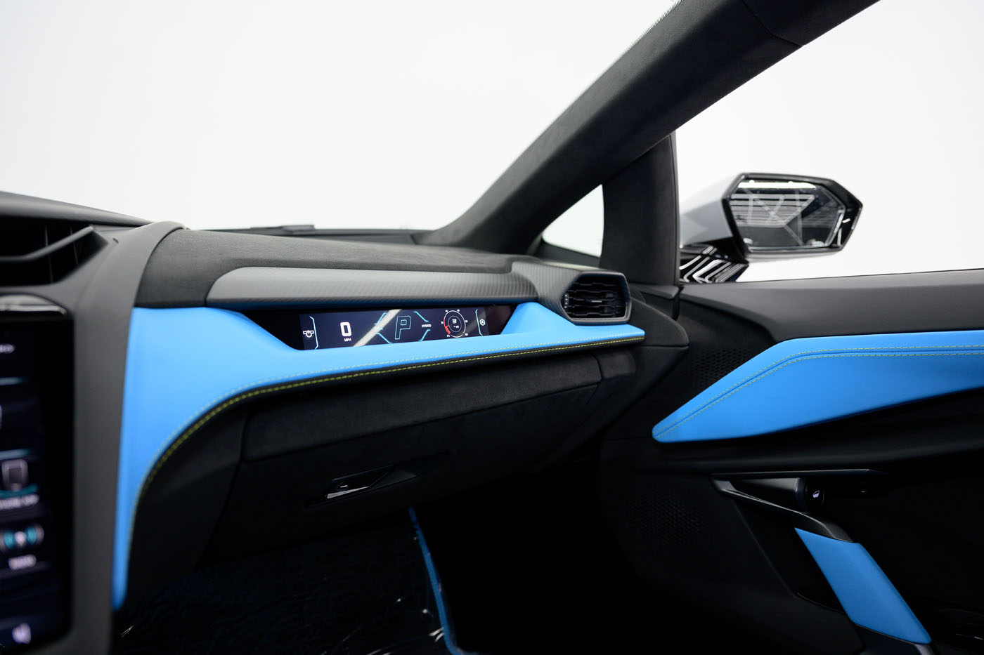 The image shows the interior passenger side of a modern car, featuring blue accents, a digital display on the dashboard, and a side mirror visible through the window—reminiscent of the luxurious 2025 Mansory Lamborghini Revuelto available in the U.S.
