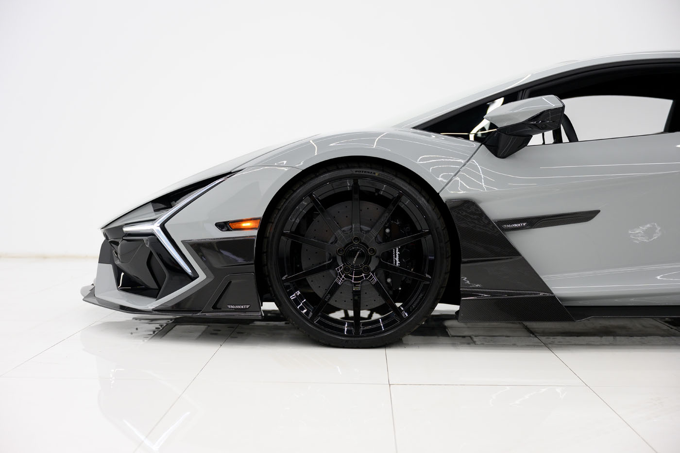 Side view of a gray 2025 Mansory Lamborghini Revuelto Limited Spec, focusing on the front wheel and aerodynamic body detailing, set against a white background in the U.S.