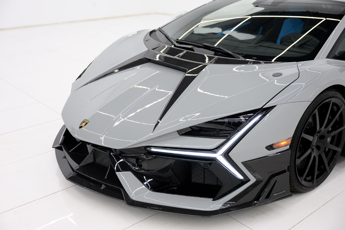 Close-up view of the front end of a grey 2025 Mansory Lamborghini Revuelto sports car with black accents, sharp angular lines, and LED headlights on a white showroom floor in the U.S.