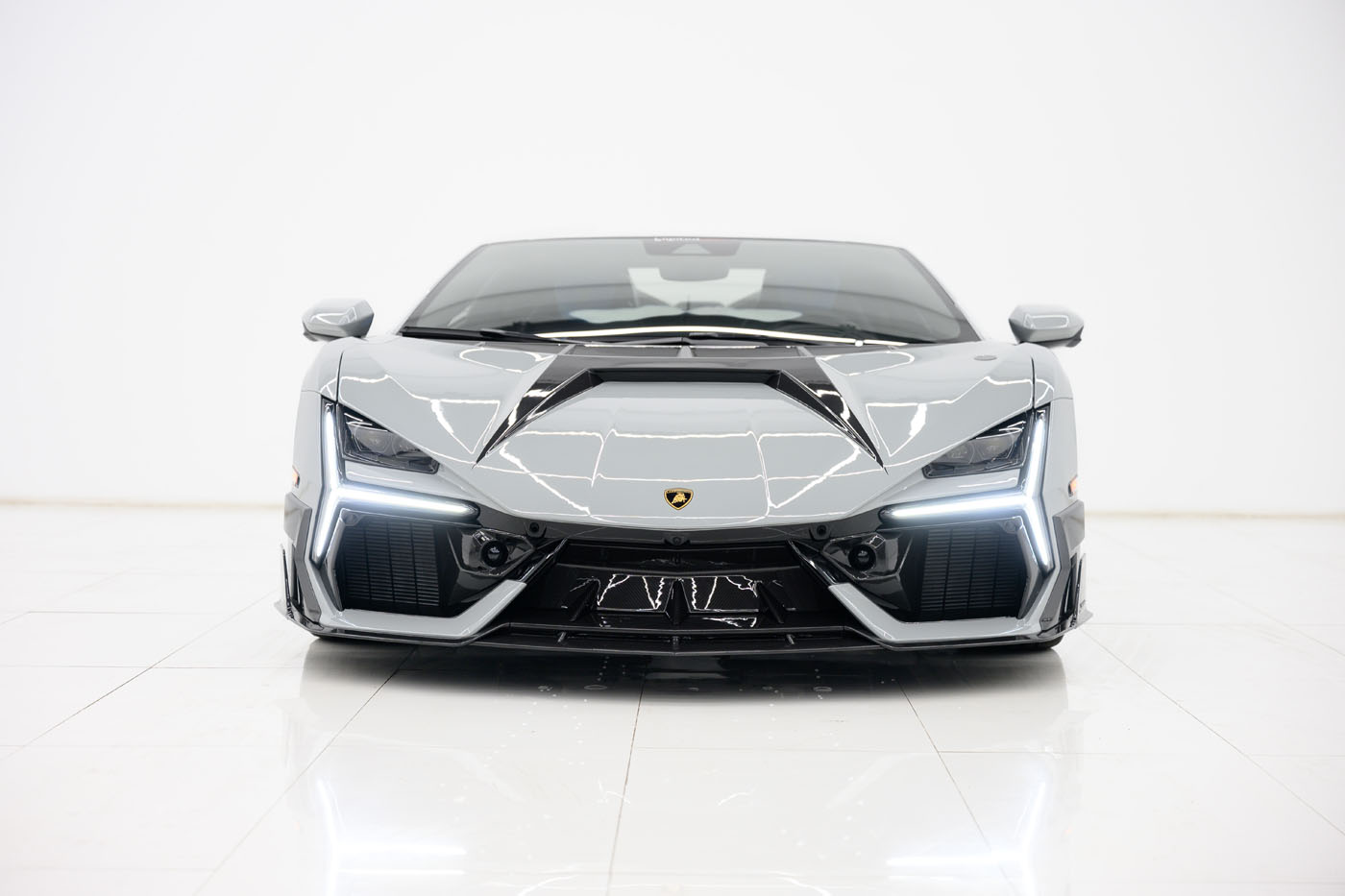 A grey 2025 Mansory Lamborghini Revuelto is photographed head-on in a bright, white U.S. showroom with its headlights on.