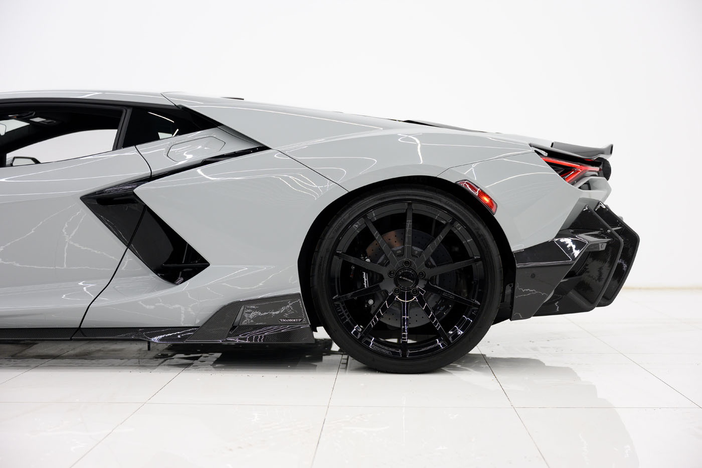 Side view of the rear half of a sleek grey 2025 Mansory Lamborghini Revuelto with black wheels, angular body lines, and a minimal white background. This exclusive model captures attention in the U.S. sports car scene.
