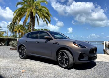 A matte gray, all-electric Maserati Grecale Folgore SUV is parked near palm trees by a waterfront in the sunny Florida Keys under a clear sky with scattered clouds.