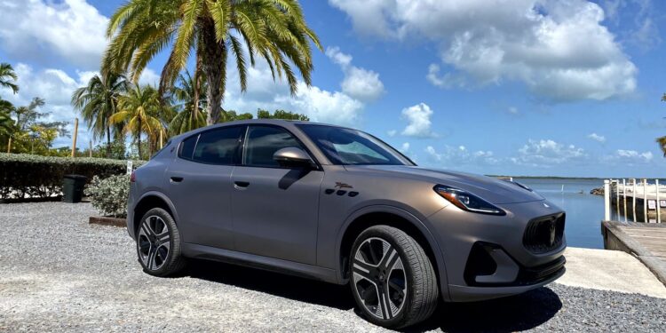 A matte gray, all-electric Maserati Grecale Folgore SUV is parked near palm trees by a waterfront in the sunny Florida Keys under a clear sky with scattered clouds.