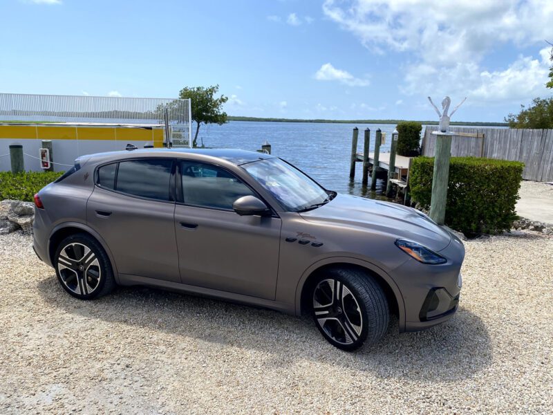 A matte gray Maserati Grecale Folgore SUV is parked on a gravel lot near water in the Florida Keys, with docks, lush greenery, and a blue sky completing the scene.