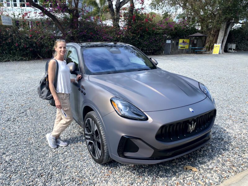 A woman stands next to a gray Maserati Grecale Folgore SUV in a gravel parking lot, surrounded by trees and flowers, capturing the all-electric luxury vibe of the Florida Keys.