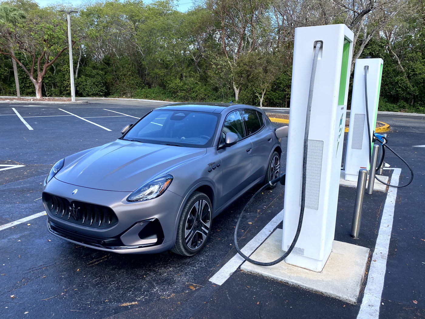 A gray Maserati Grecale Folgore, an all-electric SUV, is parked at a charging station in an empty parking lot surrounded by trees.
