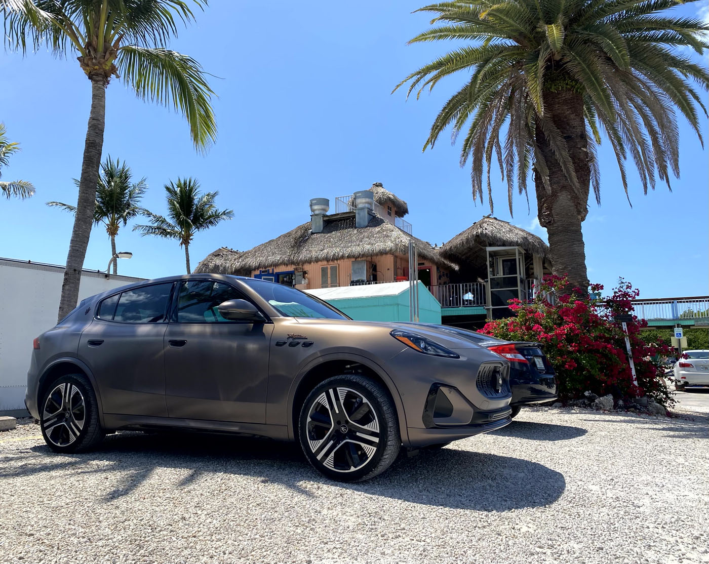 A matte-finish Maserati Grecale Folgore SUV is parked on gravel near palm trees and a thatched-roof building, all set under the clear blue sky of the Florida Keys.