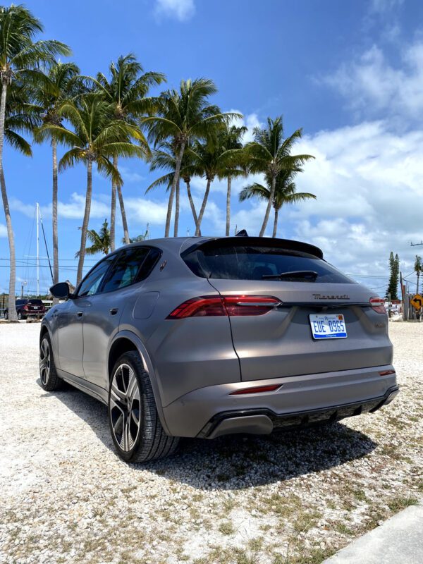 A matte gray, all-electric Maserati Grecale Folgore SUV parked on a gravel lot near palm trees under a blue sky with scattered clouds in the Florida Keys.