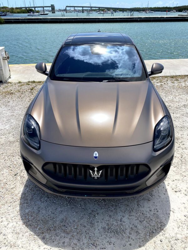 A matte bronze, all-electric Maserati Grecale Folgore SUV is parked on a gravel surface near a waterfront in the Florida Keys, with a marina and boats visible in the background.