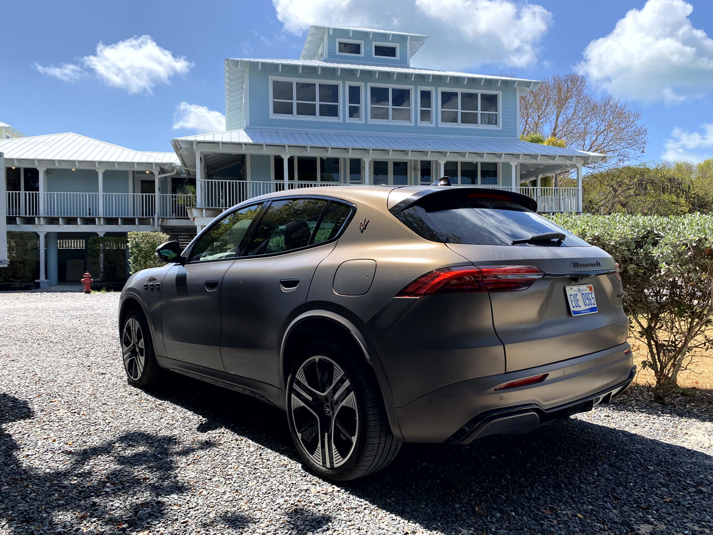 An all-electric Maserati Grecale Folgore SUV in bronze is parked on a gravel driveway in front of a large, multi-story white house with balconies.