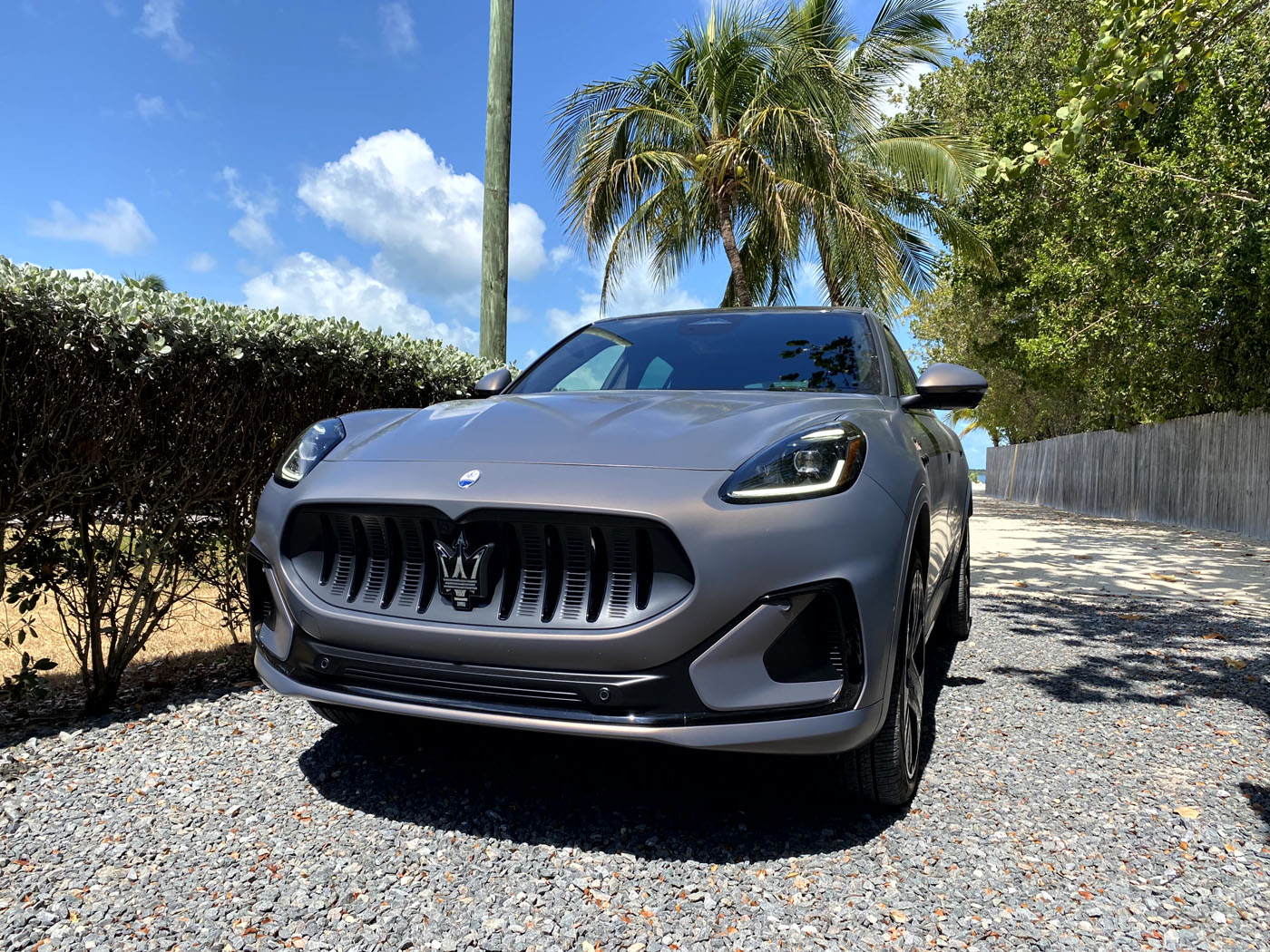 A gray all-electric Maserati Grecale Folgore SUV is parked on a gravel driveway with palm trees and a blue sky in the Florida Keys.