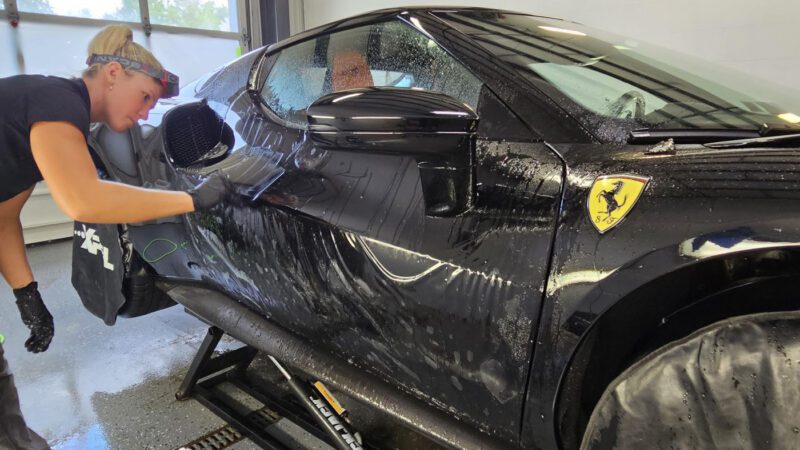A person wearing gloves gives a Ferrari 296 GTB the Royal Treatment, cleaning its side with soap and a sponge inside a WrapKings garage.