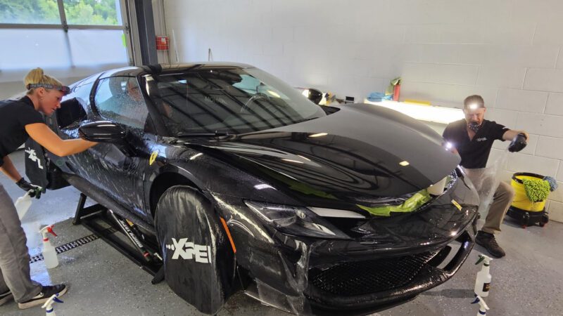 Two people from WrapKings are applying a protective film to a black Ferrari 296 GTB, giving it the Royal Treatment inside a well-lit garage.