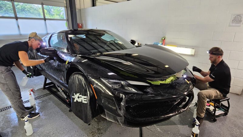 Two people apply a protective film to the exterior of a black Ferrari 296 GTB inside a well-lit garage, giving it the Royal Treatment by WrapKings.