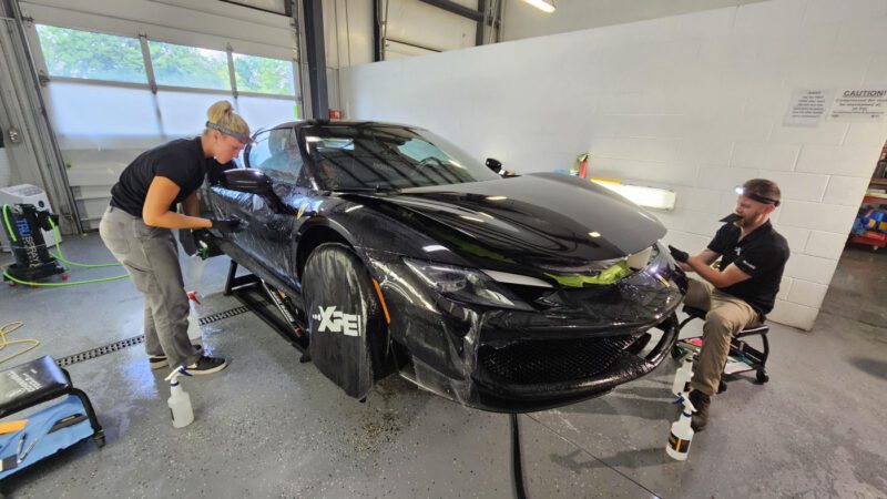 Two people are applying a protective film to the front of a Ferrari 296 GTB inside a well-lit garage, giving it the Royal Treatment by WrapKings.
