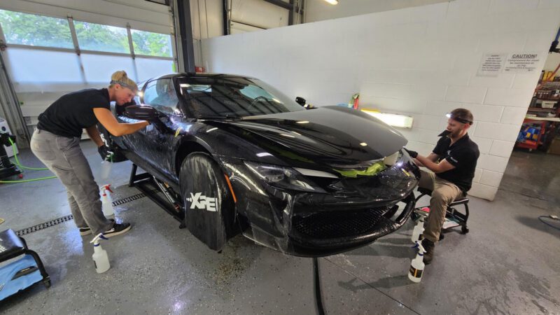 Two people give a Ferrari 296 GTB the Royal Treatment at WrapKings, carefully applying protective film with spray bottles and tools as the sleek sports car sits slightly lifted in the garage.