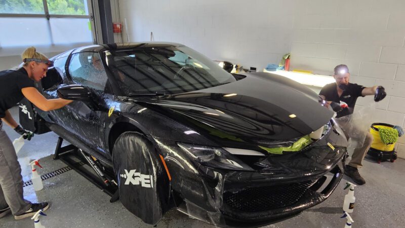 Two people give Royal Treatment to a black Ferrari 296 GTB inside a well-lit garage, carefully applying protective film with spray bottles and tools—the kind of attention to detail you expect from WrapKings.