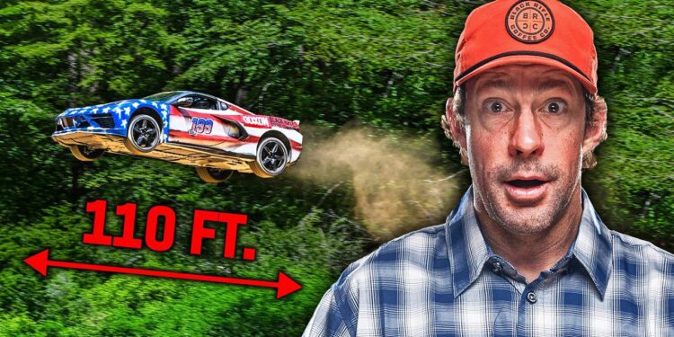 A man in a red cap stands in front of greenery as a C8 Corvette with an American flag design, driven by Travis Pastrana, soars through the air on its longest jump, marked by a "110 FT." label and arrow indicating the distance.