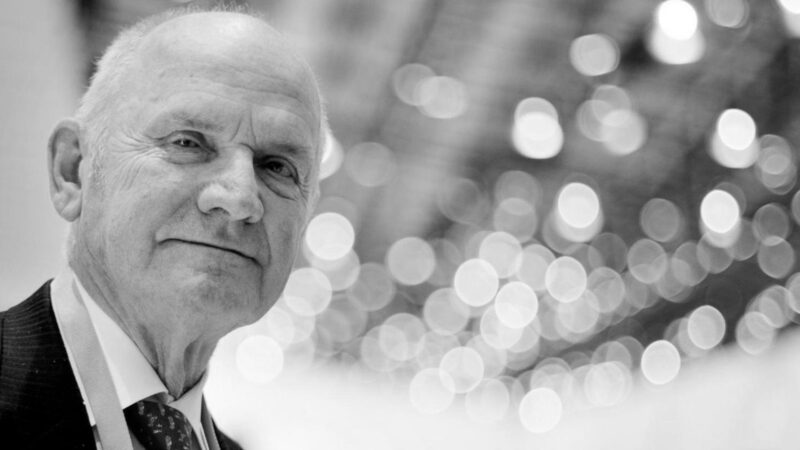 An older man in a suit and tie looks at the camera, with blurred circular lights in the background. This black and white image evokes the timeless elegance of Veyron: The Man The Machine And The Legacy That Rebuilt Bugatti.