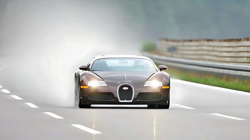 A black sports car, likely a Veyron—The Man The Machine And The Legacy That Rebuilt Bugatti—drives at high speed on a wet, empty highway, with blurred surroundings indicating motion.