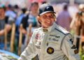 A man in a racing suit and cap smiles while standing outdoors at the Goodwood Festival, with blurred people and cameras in the background.