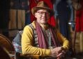 A man wearing a brown hat, red glasses, patterned vest, yellow shirt, and scarf sits in a chair indoors with his hands folded, reminiscent of the stylish champions at the Goodwood Festival.