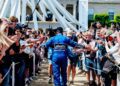 A race car driver in a blue Labatt’s Goodyear suit walks through a crowd of fans taking photos and reaching out, at the Goodwood Festival, where champions and Formula 1 legends mingle with enthusiasts.