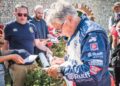 An older man in a racing suit signs autographs for fans outdoors at the Goodwood Festival, with several people holding items out for him to sign.