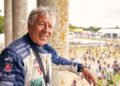 Older man in a racing suit smiles while leaning on a rusty pillar, with the excitement of the Goodwood Festival and a race track bustling with Champions in the background.