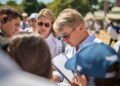 A man wearing sunglasses looks at a notepad while surrounded by a group of people outdoors at the Goodwood Festival on a sunny day.