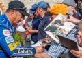 A race car driver in a blue suit, fresh from the Goodwood Festival, signs autographs for fans holding photos and memorabilia in a crowded outdoor setting.