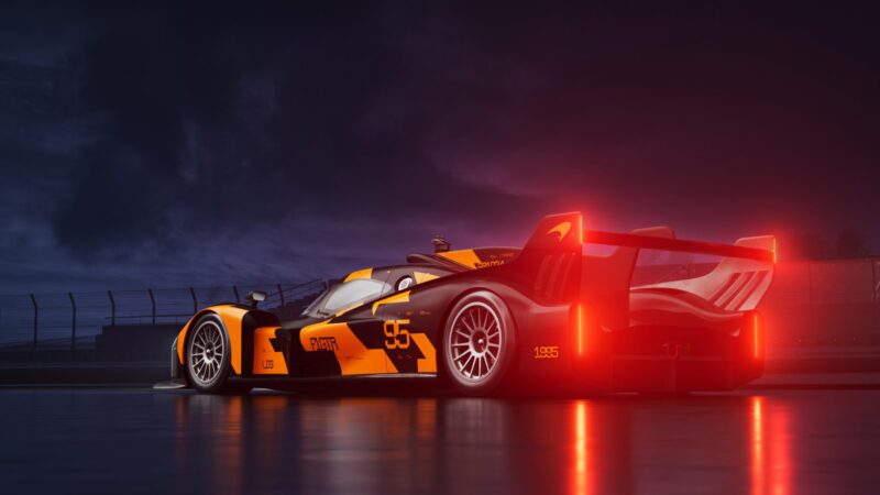 A sleek orange and black McLaren race car with illuminated tail lights is parked on a wet track at night, evoking the spirit of a Le Mans Hypercar beneath a dark, cloudy sky with a fence in the background.
