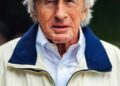 An elderly man wearing a blue and green plaid flat cap, a white collared shirt, and a cream jacket with a blue collar and "Rolex" logo—perfect attire for attending the Goodwood Festival or celebrating era-defining cars.