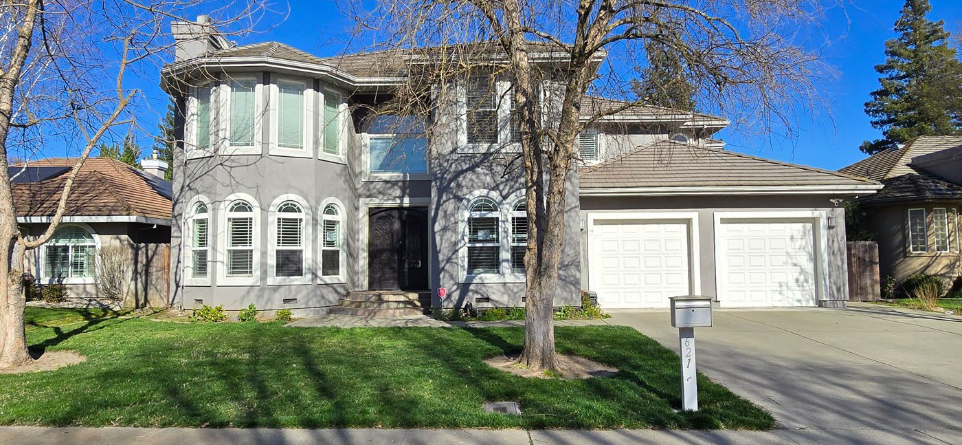 Two-story gray suburban house with white-trimmed windows, a front lawn, leafless trees, double garage doors, and a white mailbox at the curb.