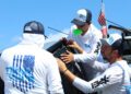 Three men wearing "Tidal Wave" shirts and hats work together on a black vehicle outdoors under a blue sky, with an American flag in the background—showcasing expert boat protection for Florida boaters.