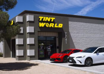 A building with a sign reading "TINT WORLD" above the entrance, with two cars—a red sports car and a white sedan—parked in front.