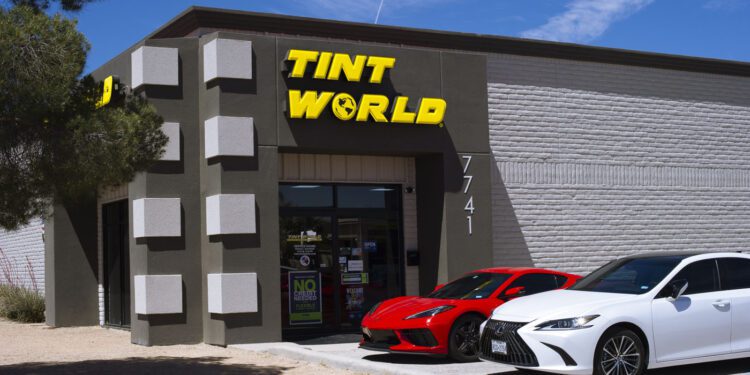 A building with a sign reading "TINT WORLD" above the entrance, with two cars—a red sports car and a white sedan—parked in front.